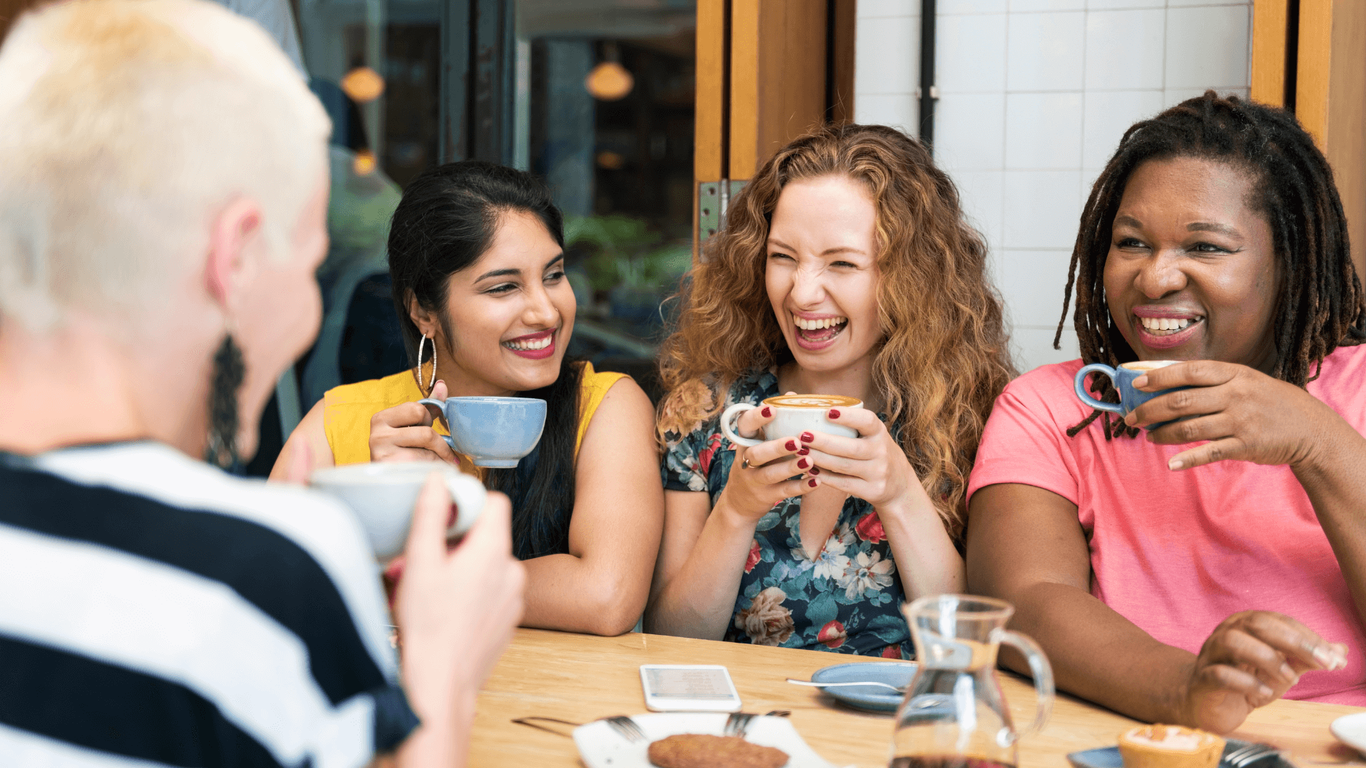 Four women of different ethnic backgrounds sit and laugh over coffee